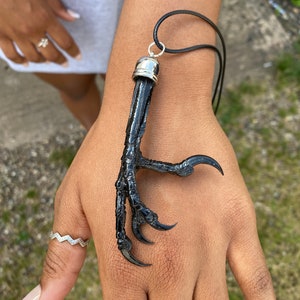 Puede incluir: Un collar con colgante de garra negra con una tapa plateada y un cordón negro. La garra tiene garras afiladas y curvadas. El collar se muestra en una mano con anillos.