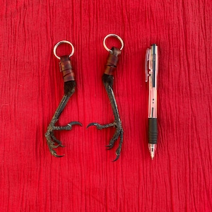 May include: Two brown leather keychains with black bird feet attached. The keychains are hanging against a red fabric background. A silver pen is on the right side of the image.