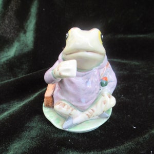 May include: A ceramic figurine of a frog wearing a purple robe and holding a cup. The frog is sitting on a green lily pad.