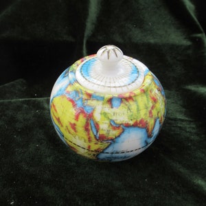 May include: A spherical ceramic jar with a lid, decorated with a world map design. The jar features a white lid with a gold-coloured starburst pattern and a small knob. The globe design includes yellow, blue, and red continents.