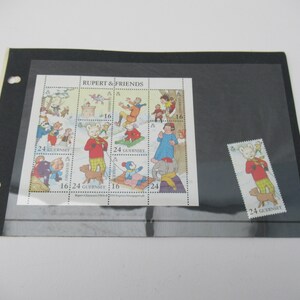 May include: A collection of Rupert & Friends stamps and a single stamp are presented in a black album page. The stamps showcase vibrant illustrations of Rupert Bear and friends. The stamps are marked "Guernsey" and have different values.
