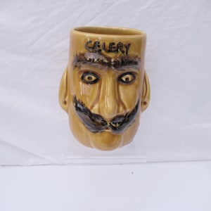 May include: A tan ceramic mug with a face design. The mug features a face with a mustache, eyebrows, and the word "CELERY" on the rim. The mug has a handle shaped like an ear.