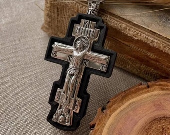 May include: A silver-colored religious cross pendant with a black border. The cross features a detailed depiction of Jesus Christ on the cross, with intricate carvings and inscriptions. The pendant is likely made of metal and is designed to be worn as a necklace.