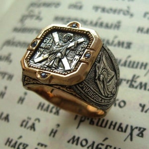 Peut inclure: Une bague en or et argent avec une icône religieuse et des pierres précieuses bleues. La bague est posée sur un livre avec du texte cyrillique.