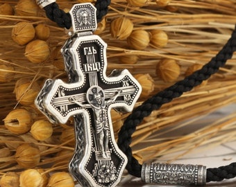 May include: A silver Orthodox cross pendant with intricate details and black enamel. The cross features a depiction of Jesus Christ on the crucifix and Cyrillic lettering. The pendant is attached to a black braided cord.