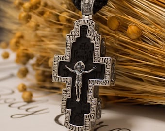 May include: A silver Orthodox cross pendant with black wood inlay and intricate engravings in Cyrillic script. The cross features a depiction of Jesus Christ on the crucifix.