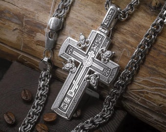 May include: A silver cross pendant with intricate details and engraved text, hanging on a silver chain. The cross features a central design with a smaller cross and the words "IC XC" and "NIKA".