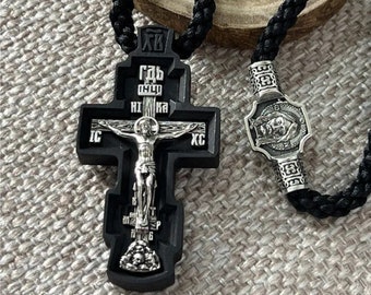 May include: A black Orthodox cross pendant with a silver crucifix and skull base, strung on a black cord. The cross features Cyrillic inscriptions and is paired with a silver bead with a religious image. The background is a textured, neutral fabric.
