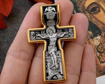 May include: A silver and gold cross pendant with a black background. The cross features a depiction of Jesus Christ on the cross, with figures of angels and saints on the sides. The cross is adorned with intricate details and a skull and crossbones at the base.