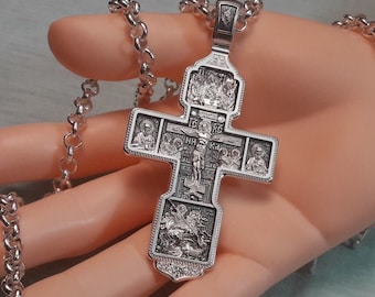 May include: A detailed silver pendant cross with intricate religious iconography, including figures and scenes. The cross is suspended from a matching silver chain. The design features detailed engravings and a polished finish, showcasing a traditional style.