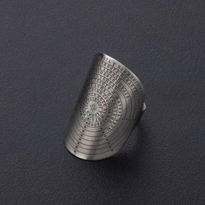 May include: A silver-toned ring with an engraved design featuring concentric circles of text. The ring has a wide band and a curved face, with the text appearing to be in a foreign language. The ring is set against a dark background.