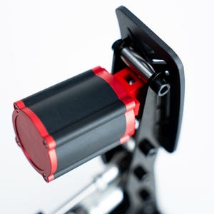Puede incluir: Primer plano de un componente mecánico negro y rojo. El cuerpo cilíndrico es negro con detalles rojos, montado sobre un soporte negro. El diseño sugiere un instrumento de precisión o parte de una máquina más grande.