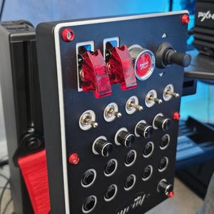 May include: Black control panel with red detailing, including toggle switches, rotary knobs, and an "ENGINE START" button. The panel is mounted on a red and black frame, possibly for a racing simulator.