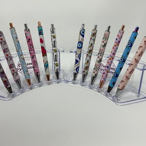 May include: A collection of ballpoint pens with various designs, including Hello Kitty, butterflies, and an evil eye. The pens are displayed in a clear, tiered holder. Some pens have the word "SENNA" printed on them.