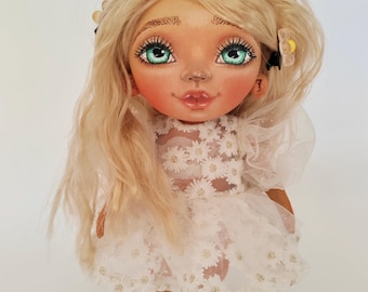 Könnte beinhalten: Eine handgefertigte Puppe mit langem blondem Haar, grünen Augen und rosigen Wangen. Die Puppe trägt ein weißes Kleid mit Gänseblümchen-Verzierungen und passenden Schuhen. Die Puppe hat einen süßen Ausdruck und ist mit kleinen Haaraccessoires geschmückt.