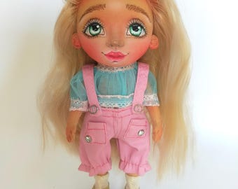 Könnte beinhalten: Handgefertigte Puppe mit langem blondem Haar und grünen Augen. Die Puppe trägt ein blaues Oberteil mit Spitzenbesatz, eine rosa Latzhose mit Strassdetails und gelbe Schuhe. Kleine Haarspangen mit Blumen schmücken das Haar der Puppe.