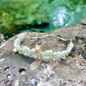 May include: A delicate silver bracelet with green and white beads and small seashells. The bracelet is lying on a gray rock.