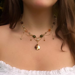 Puede incluir: Un collar dorado con un colgante de hoja. El collar presenta una delicada cadena con pequeñas cuentas en tonos verdes, marrones y naranjas. Delicadas cadenas doradas cuelgan del collar. El colgante es una hoja dorada.