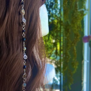 May include: A silver hair chain with black beads, white skull beads, and silver swirl beads. The chain is hanging in a woman's brown hair.