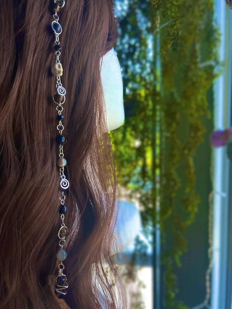 May include: A silver hair chain with black beads, white skull beads, and silver swirl beads. The chain is hanging in a woman's brown hair.