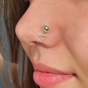 Puede incluir: Un aro de nariz de oro con una piedra preciosa verde. El aro está hecho de alambre que está retorcido en forma de espiral.