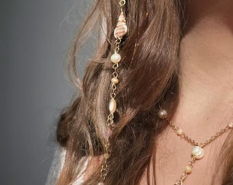 May include: A gold chain hair accessory with small seashells and pearls. The chain is hanging down a woman's brown hair.