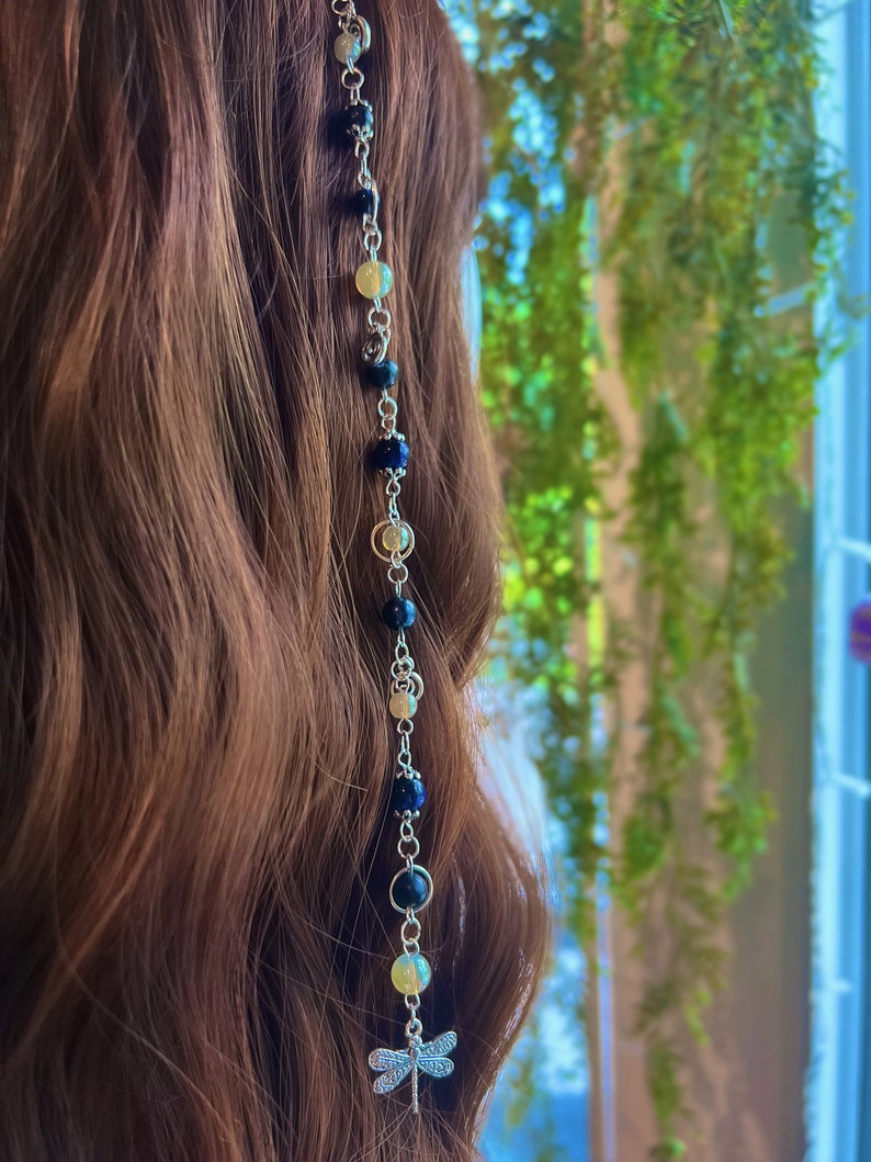 May include: A silver chain with blue and yellow beads and a dragonfly charm. The chain is hanging from a person's hair.