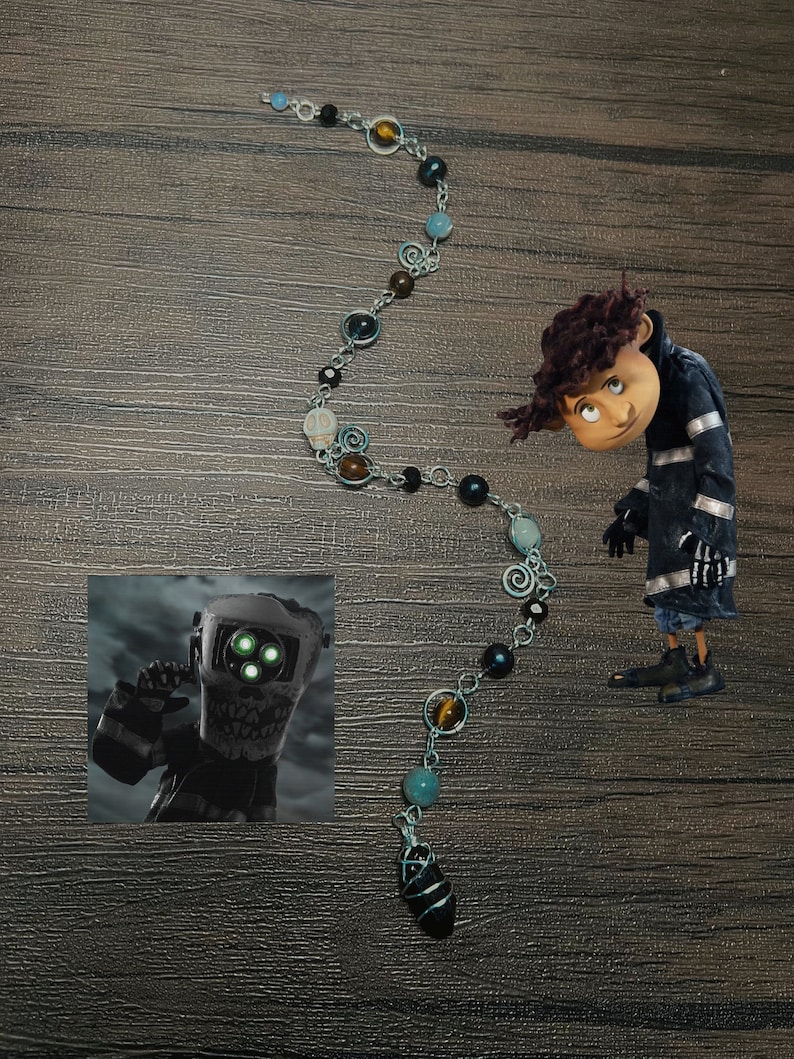 May include: A silver chain necklace with a black stone pendant. The chain features various beads, including black, brown, blue, and green. The necklace is on a wooden surface.