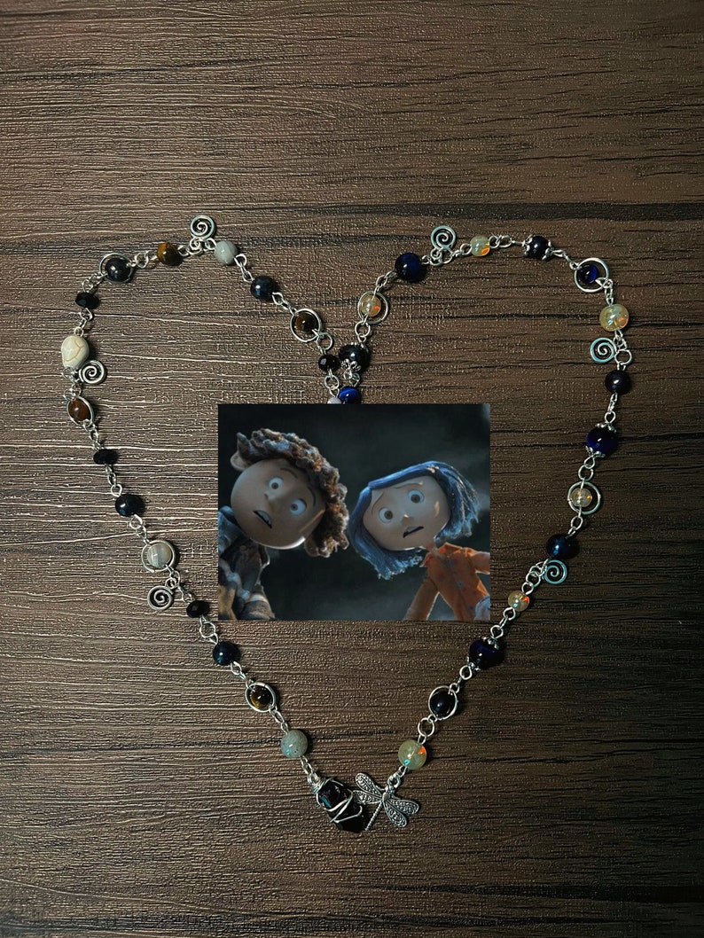 May include: A silver necklace with a dragonfly charm and various colored beads, including black, brown, blue, and white. The necklace is shaped into a heart and a picture of two characters from the movie "Coraline" is in the center.
