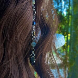 May include: A silver chain with a variety of beads, including black, brown, and green, hangs from a person's brown hair. The chain ends with a large, dark, faceted crystal.