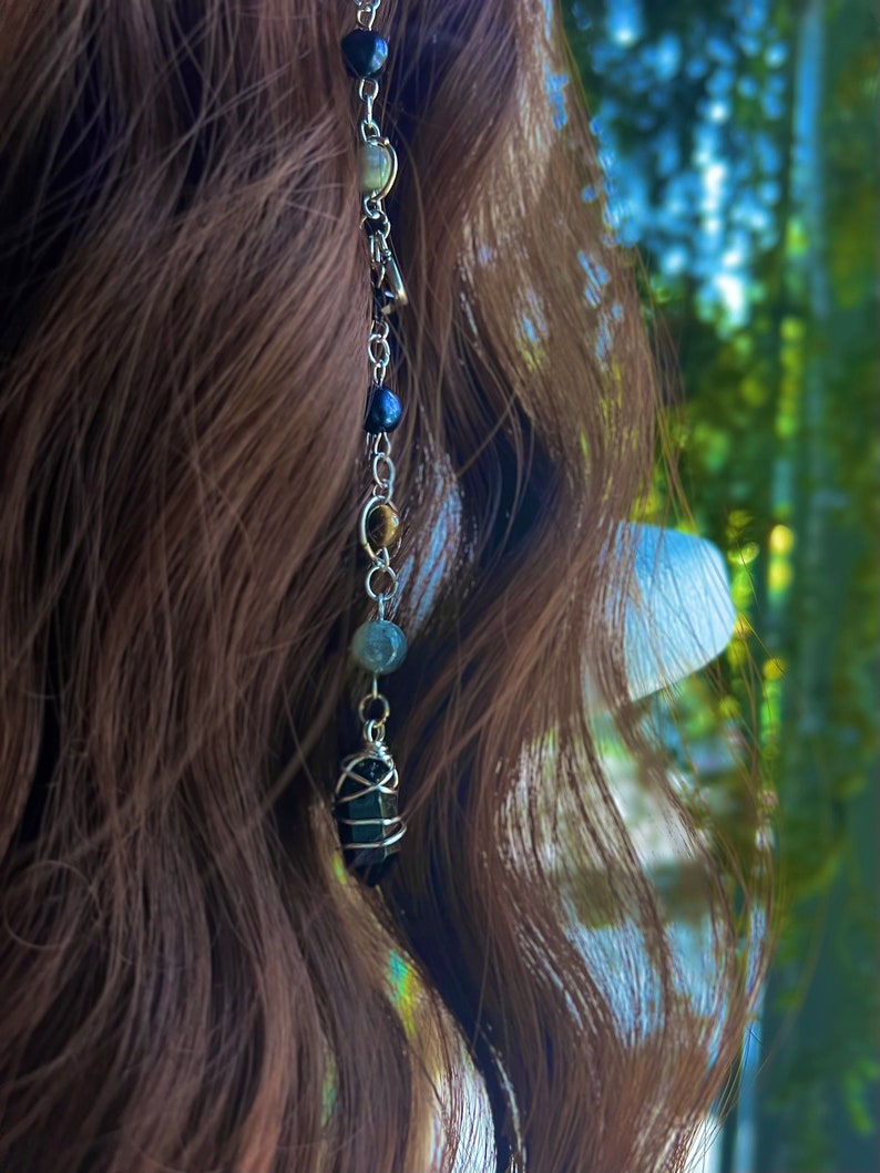 May include: A silver chain with a variety of beads, including black, brown, and green, hangs from a person's brown hair. The chain ends with a large, dark, faceted crystal.