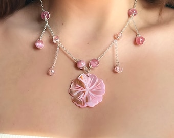Puede incluir: Un delicado collar de cadena plateada con cuentas de vidrio rosa y un gran colgante de flor rosa. El colgante de flor tiene un diseño detallado y un acabado nacarado.