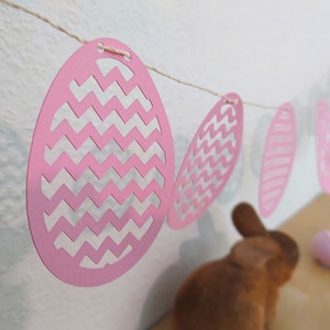 Può includere: Una ghirlanda di carta con motivo a chevron rosa e bianco con uova di carta rosa appese a uno spago bianco. La ghirlanda è appesa a un muro bianco.