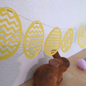 Può includere: Uno striscione di uova di Pasqua di carta giallo e bianco con diversi motivi, tra cui zig zag, strisce e pois. Lo striscione è appeso a un muro bianco. Un coniglio di peluche marrone è seduto su un tavolo sotto lo striscione.