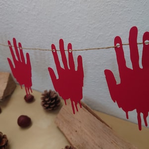 May include: A red paper Halloween garland with five hanging paper hands dripping with fake blood. The garland is strung with twine and is hanging against a white wall.