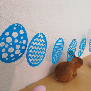 Può includere: Una ghirlanda di uova di Pasqua di carta blu e bianca con diversi motivi su ogni uovo. La ghirlanda è appesa a un muro bianco e in primo piano c'è una statuetta di coniglio marrone.