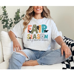 May include: A light gray short-sleeve t-shirt featuring the text FAIR SEASON IS MY FAVORITE SEASON in colorful, distressed block letters. The design includes silhouettes of a chicken, a cow, a prize ribbon, and a Ferris wheel graphic.