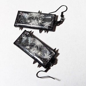 May include: A pair of black earrings with silver glitter and white star designs. The earrings are rectangular in shape and are attached to a black hook.