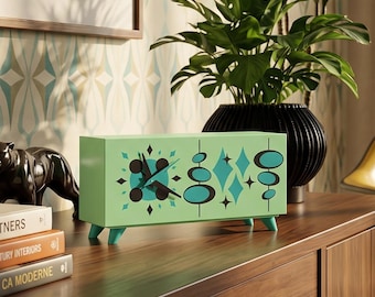 May include: A rectangular mint green desk clock featuring a black clock face with starburst and oval motifs in black and teal. The clock stands on four small legs and is positioned on a wooden surface next to books and a black panther figurine.