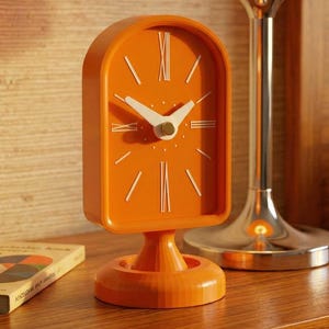 Peut inclure: Une horloge orange de style rétro avec un cadran et des aiguilles blancs. L'horloge est posée sur un petit socle. Une lampe avec un abat-jour orange est à l'arrière-plan. Un livre avec une couverture colorée est à gauche.