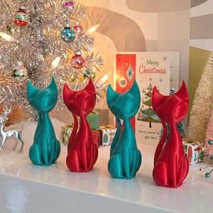 May include: A festive display featuring a silver tinsel Christmas tree adorned with ornaments and lights. Four cat figurines in teal and red are arranged on a white shelf, alongside decorative trees, deer figurines, and a Christmas card.