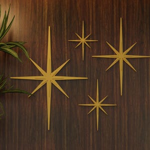 May include: Four gold starburst wall decorations against a dark wood background. The starbursts vary in size and have pointed rays. A green plant with long leaves is in the upper left corner.