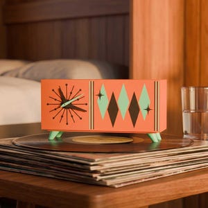 May include: A rectangular orange clock with a sunburst dial on the left and a pattern of green and dark brown diamonds on the right. The clock features light green feet and is positioned on top of a stack of black vinyl records.