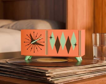 May include: A rectangular orange clock with a sunburst dial on the left and a pattern of green and dark brown diamonds on the right. The clock features light green feet and is positioned on top of a stack of black vinyl records.