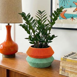 May include: A modern, two-toned planter in orange and green holds a lush green plant. The planter sits on a wooden cabinet next to a vintage orange lamp and a stack of magazines. A framed mid-century modern art print hangs on the wall.