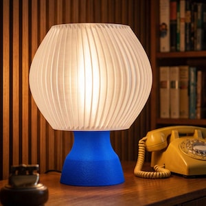 May include: A table lamp with a bright blue, textured, flared base and a translucent, white, pleated, spherical shade that emits a soft, warm glow. The lamp is positioned on a wooden surface next to a yellow rotary telephone and a small metal object.