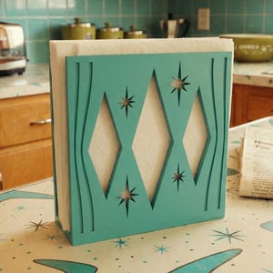 May include: A teal napkin holder featuring a cutout design of three vertical diamonds and four-pointed starbursts. The flat, rectangular structure is constructed from a solid material and holds a stack of white paper napkins in an upright position.