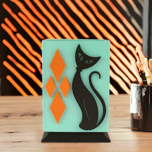 May include: A teal blue ceramic holder with a black silhouette of a cat and four orange diamond shapes. The holder has a black base.