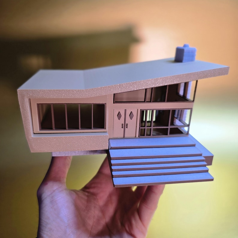 May include: A beige and brown model home with a flat roof and a chimney. The home has a large window on the front and a set of double doors. The home is sitting on a set of blue stairs.