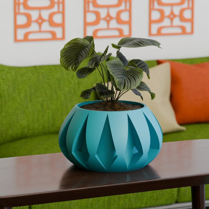 May include: A teal-colored, geometric planter with a leafy green plant inside. The planter has a unique design with diamond-shaped cutouts. The plant pot sits on a dark brown table, with a green couch and orange wall decor in the background.